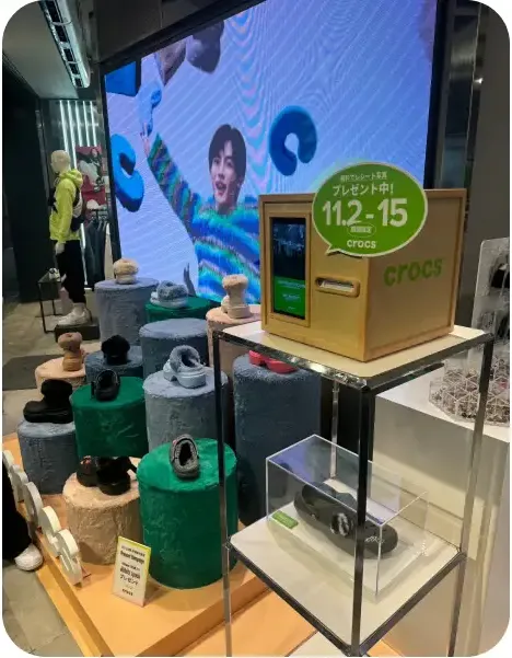 매장 진열대 위 영수증사진기와 crocs 브랜드 신발 전시, 대형 스크린 광고